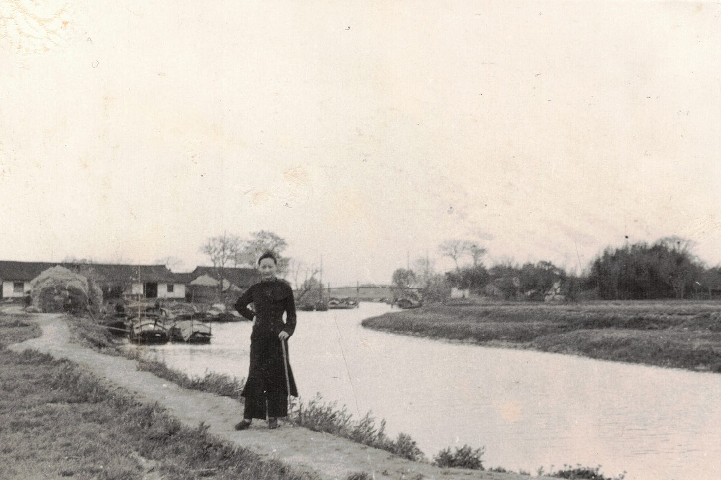 Anna May Wong in all black stands on a dirt path near a canal with farmhouses in the background, rural China