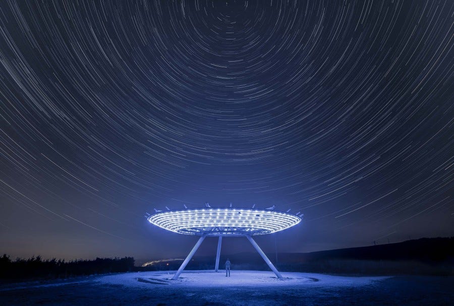 A long-exposure photo shows star trails forming circles above an illuminated sculpture on a hill.
