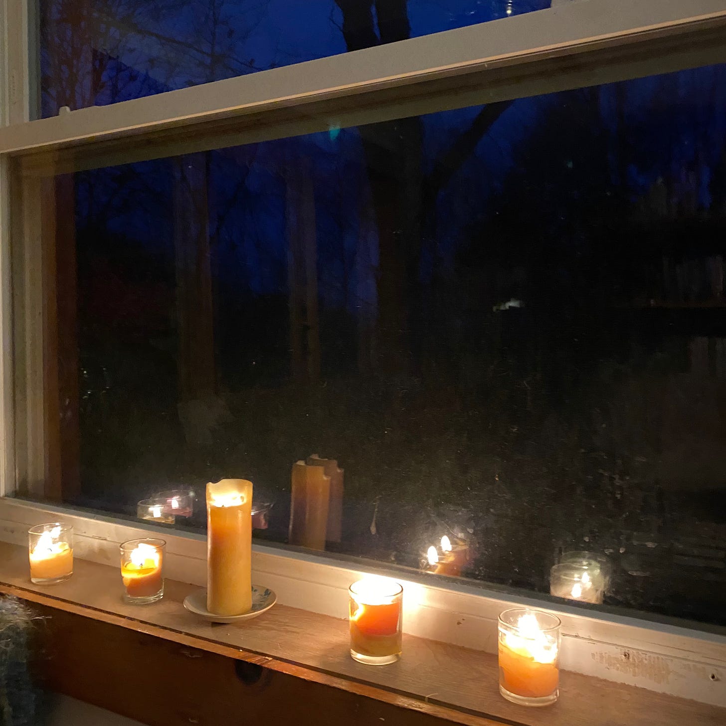 Five small lit candles on a windowsill. The sky outside the window is a deep, dark, pre-dawn blue. A few black tree branches are visible.