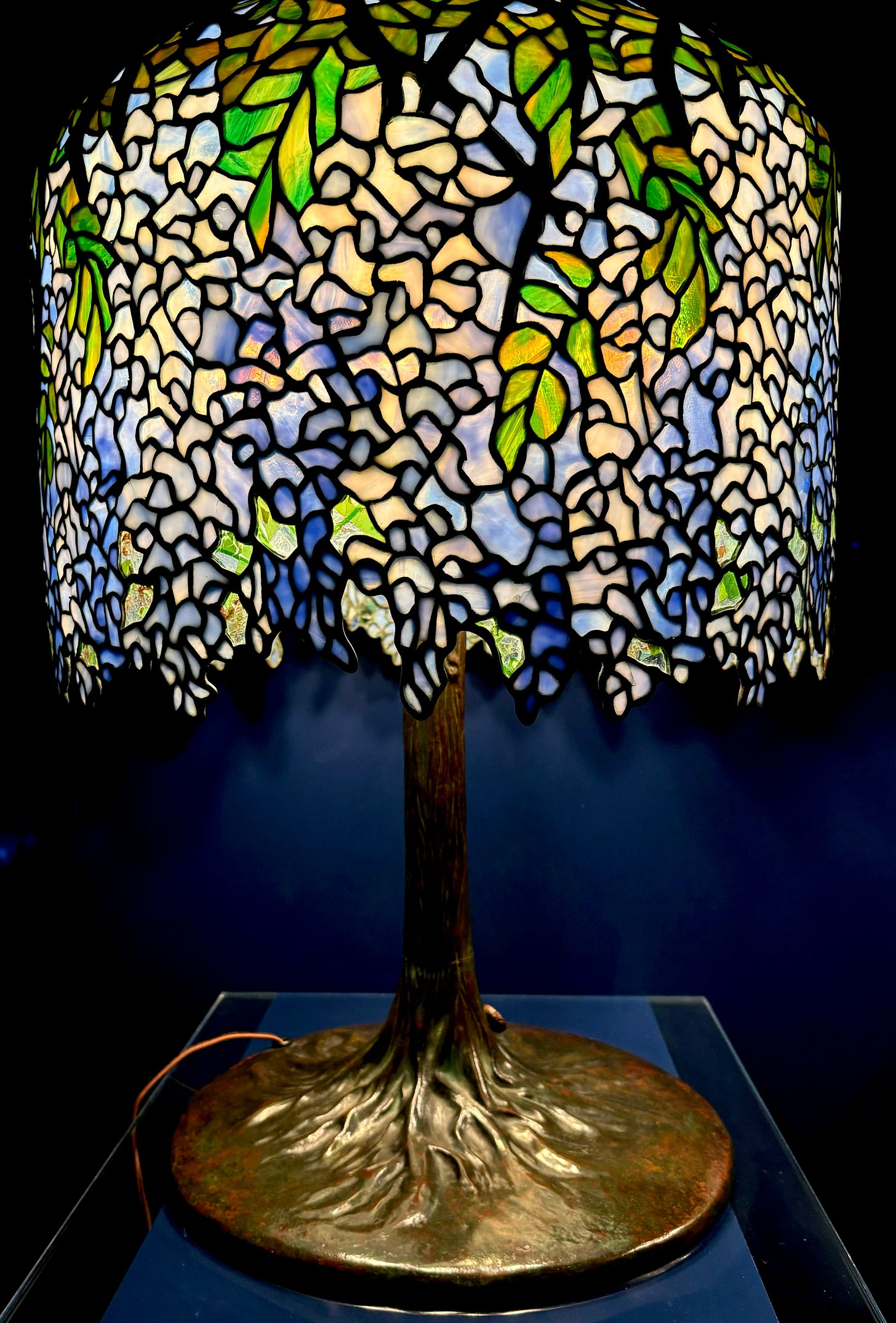 A wisteria lamp. Different pieces of pale blue glass come together to form wisteria petals, with green glass forming leaves on the top of the lamp shade. The base is metal that looks almost like lustrous wood.