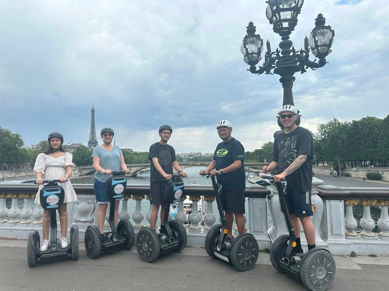 A photograph of a family of five people, all on segways, wearing helmets, on a bridge in Paris. You can see the Eiffle Tower behind them in the distance. They're all smiling, looking at the camera. 