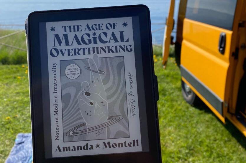 In the foreground, I hold up my Kindle showing the cover of Amanda Montell's Age of Magical Overthinking. In the background, our yellow converted campervan sits in a grassy campsite by the water