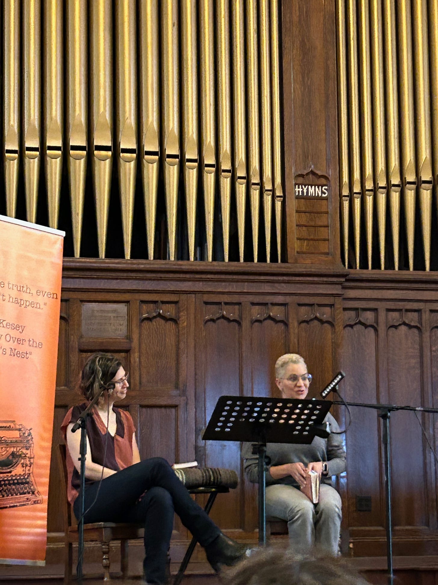 Image shows author Idra Novey and author Asale Angel-Ajani discussing their books on a church stage beneath organ pipes