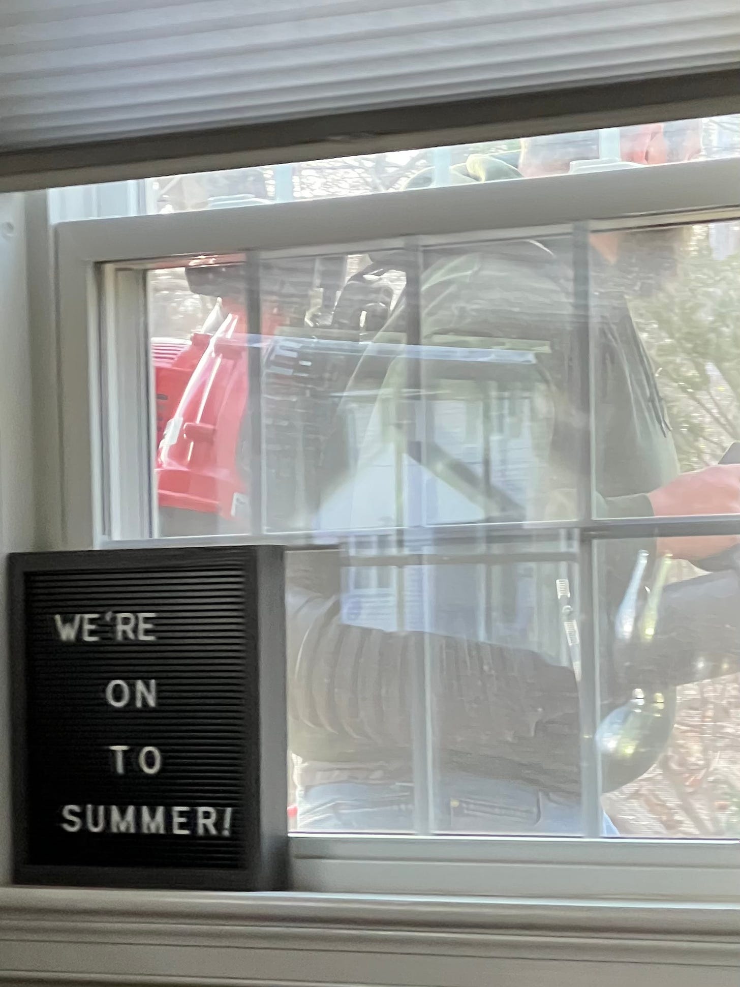 Picture of a man with a leaf blower right outside a window; a sign in the window reads “We’re on to summer”