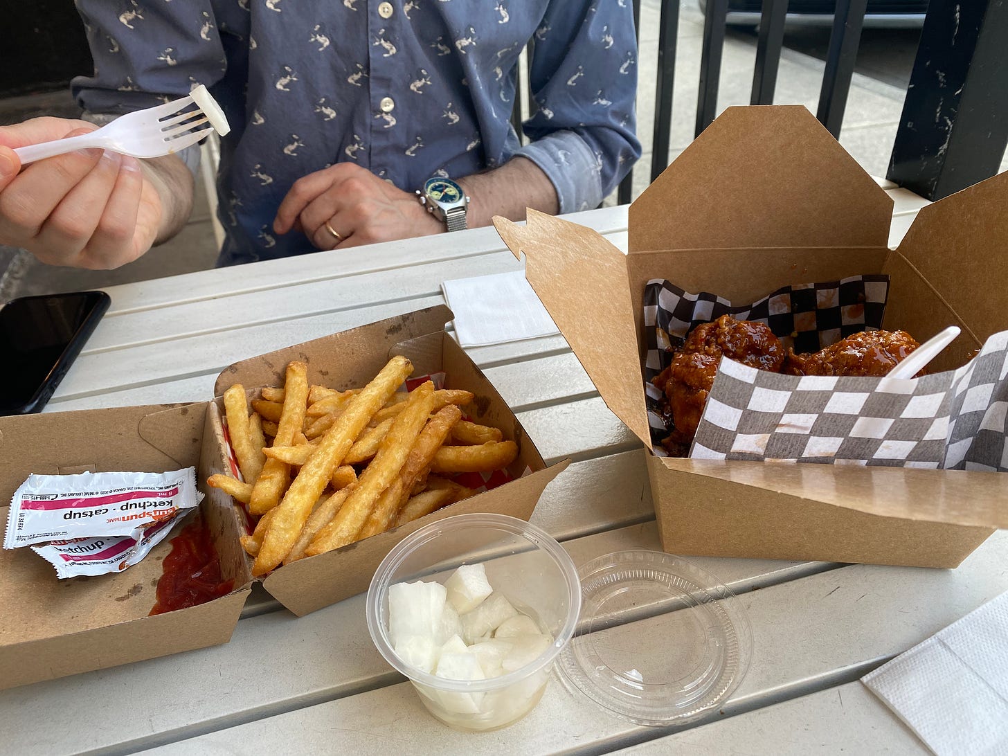An outdoor table with paper containers of fries and chicken. Ketchup has been squeezed into the container beside the fries, the empty packets next to it. In front of the two containers is a small plastic ramekin of pickled radish. In the background, Jeff holds a plastic fork with a piece of radish on it. 