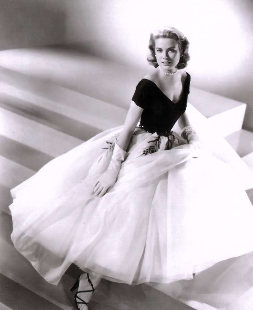 A black-and-white photo of Grace Kelly sitting dramatically on some stairs in a dramatic fluffy dress and strappy heels.
