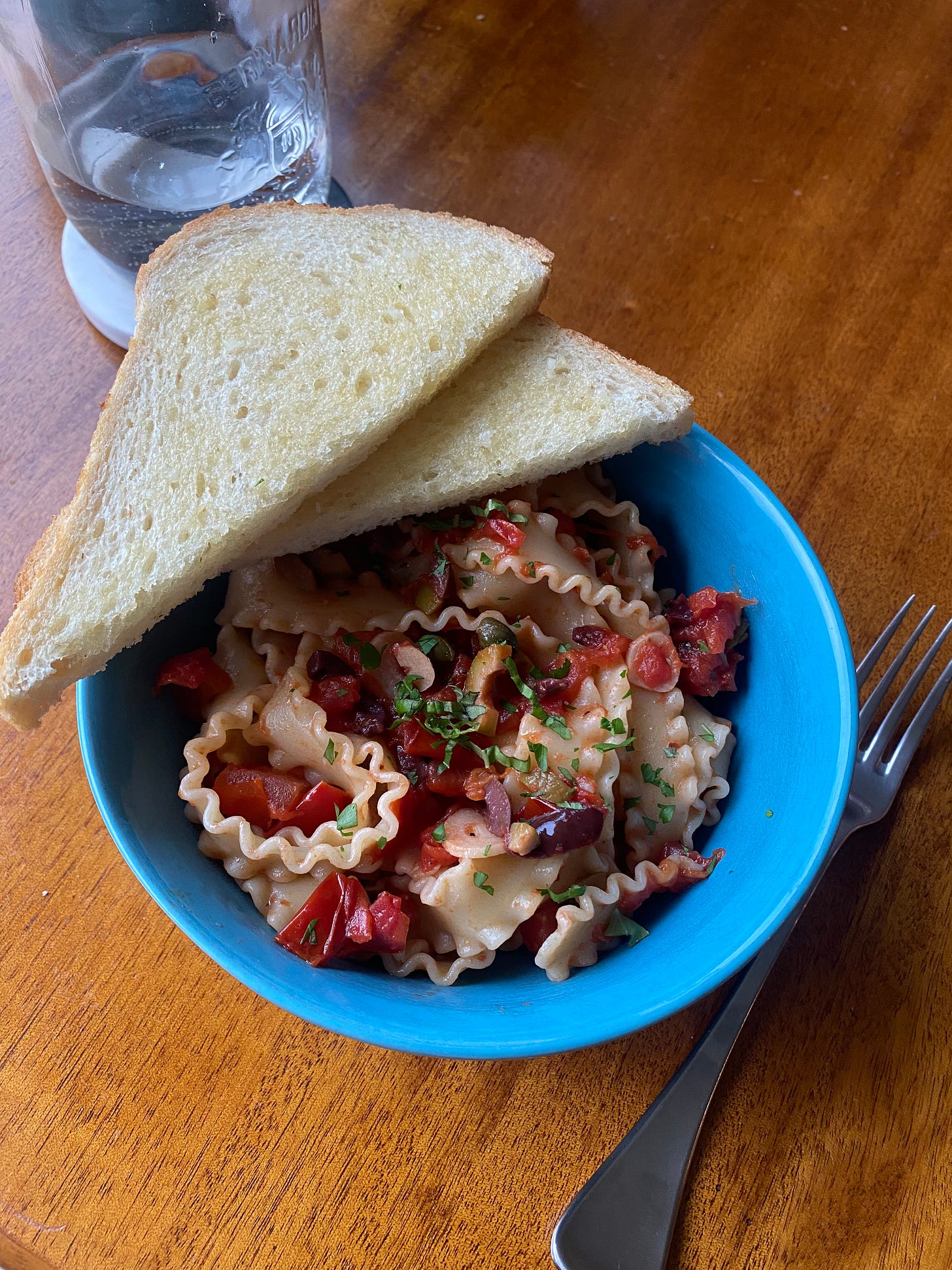 a blue bowl of pasta, thick wavy noodles, in puttanesca sauce. two halves of buttery sourdough rest at the edge of the bowl.