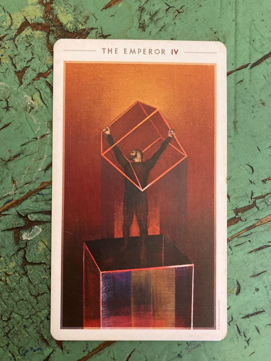 The Emperor tarot card lies on a green wooden surface. The card depicts a strong man standing on a cube, holding up a cube that fits over the top half of his body