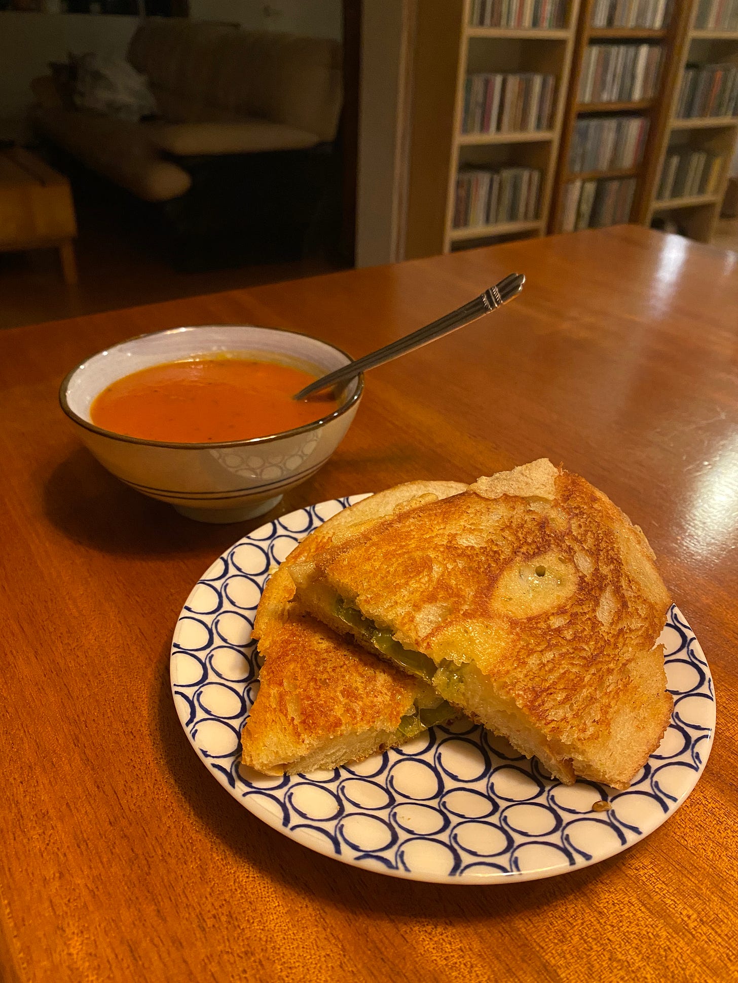 A grilled cheese sandwich with shishito peppers on white sourdough, cut in half on a small plate. Behind it is a little bowl of tomato soup with a spoon sticking out of it.