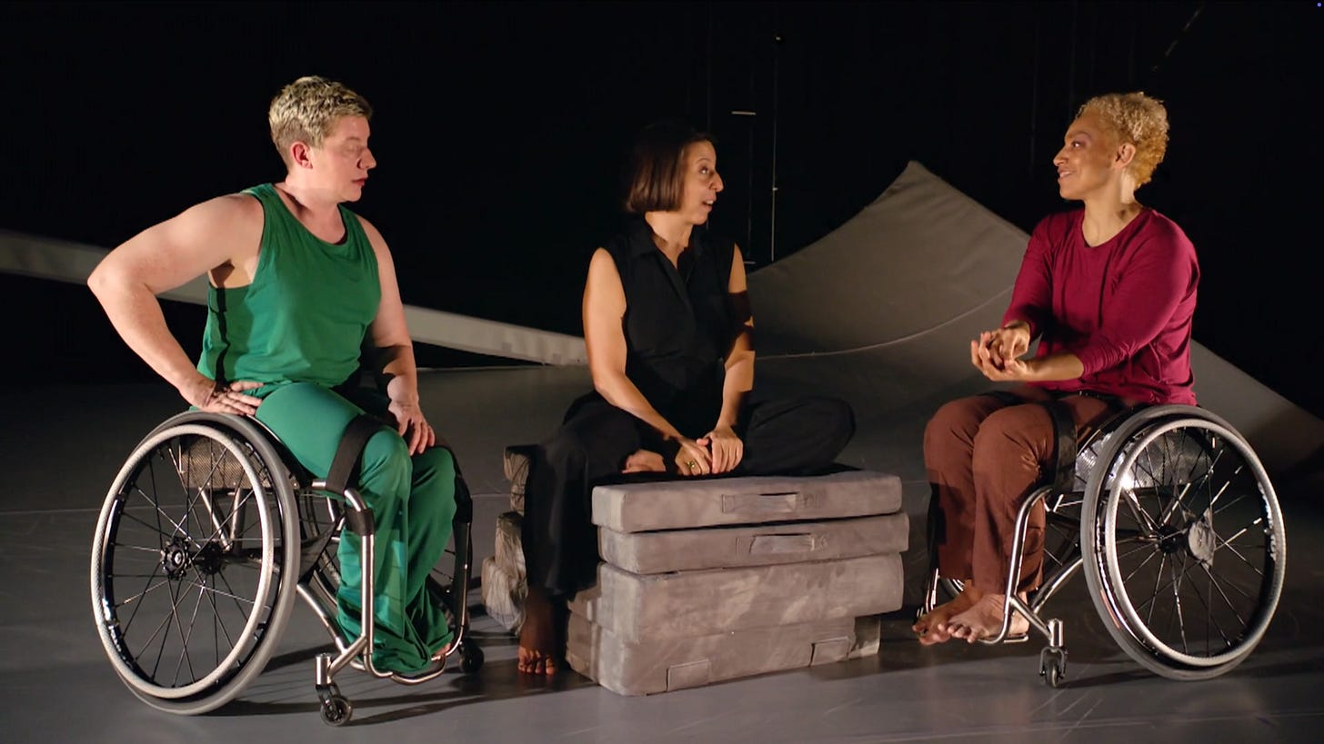 A nondisabled white woman sits on cushions between two artists in manual wheelchairs. One is a white person with pale freckled skin and very short cropped hair wearing a green sleeveless top and matching pants. A Black multiracial woman with blonde curly hair wearing a maroon long sleeve top and brown pants is smiling as she talks.