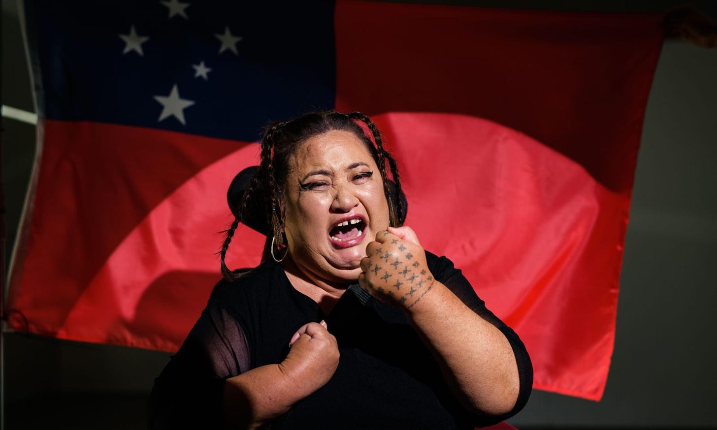 A disabled femme using a powerchair is mid-cry under a spotlight with raised fists against the Samoan flag.