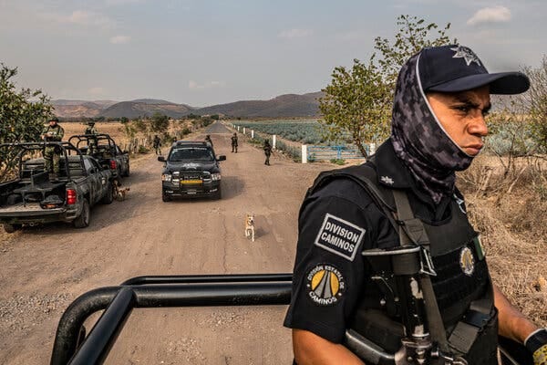 A Mexican police officer rides atop a vehicle as it passes through a checkpoint where military vehicles are parked along the side of the dirt road. 