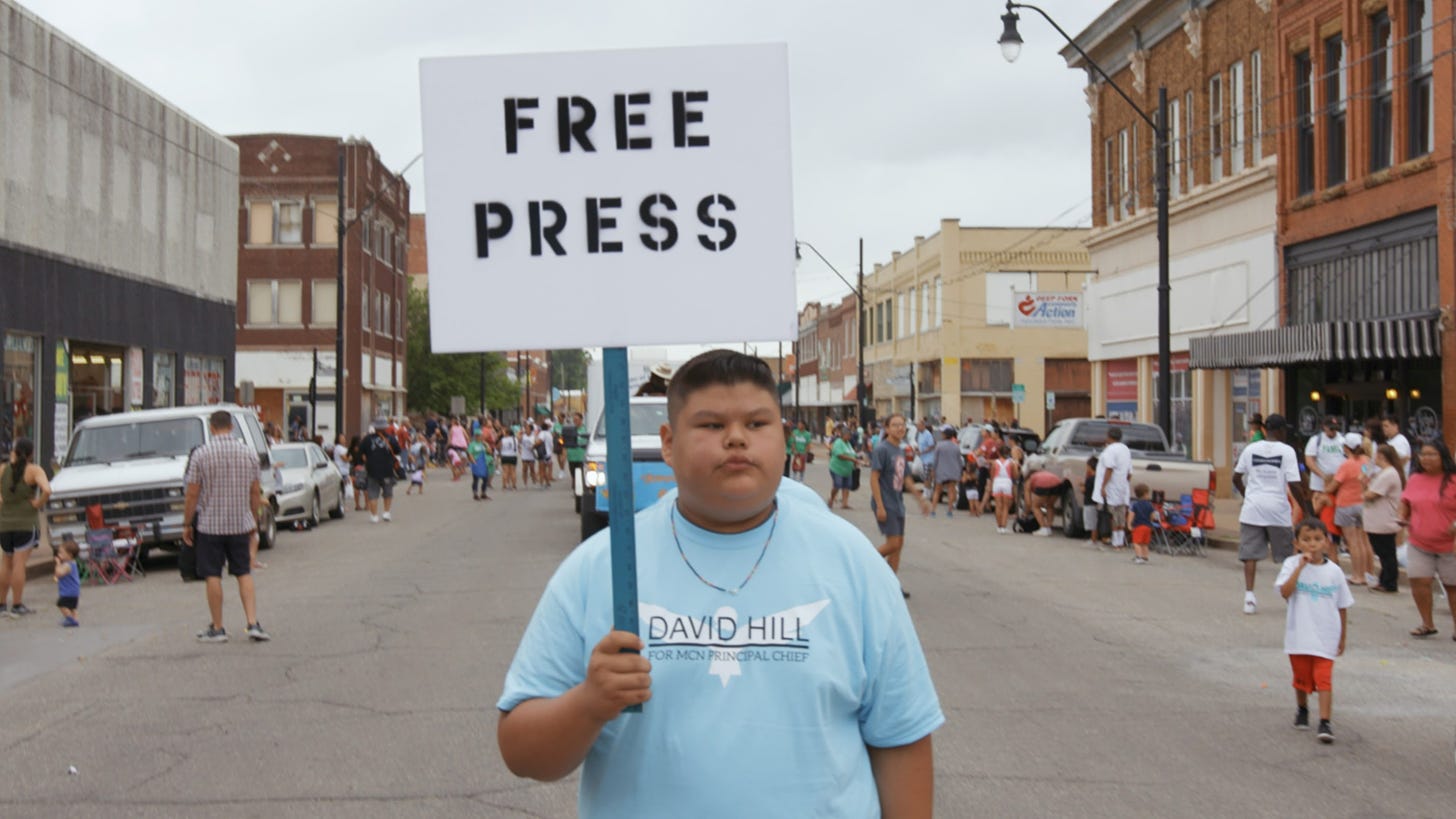 Young Muskogee boy holds sign that reads "Bad Press" in a crowded street or parade
