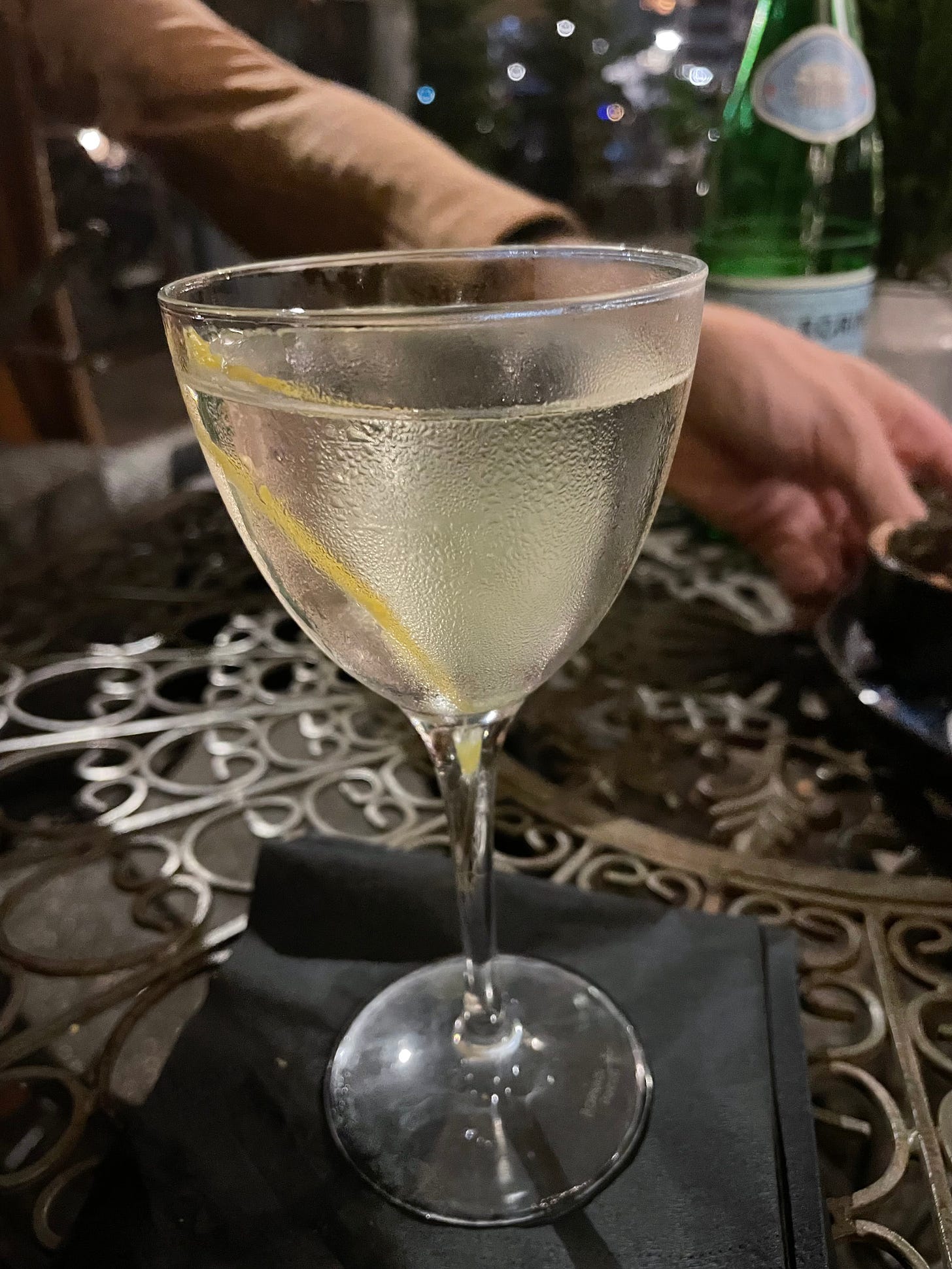 gin martini in a coupe glass with a lemon twist, on a wrought-iron table. A hand of a fellow diner and a bottle of sparkling water in the background.