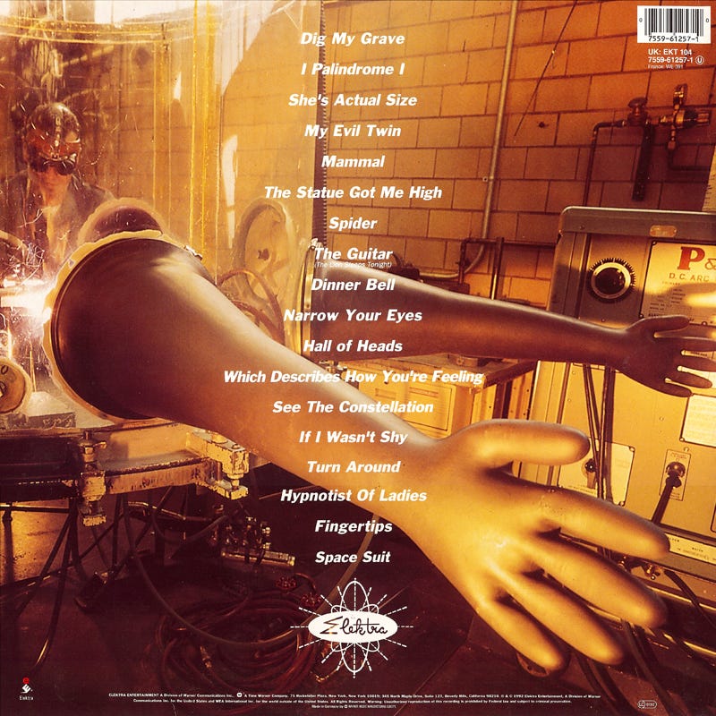 Back cover of They Might Be Giants’ Apollo 18 album, featuring a surreal lab scene with oversized mannequin arms inside a glass chamber, industrial pipes, and a song list including “If I Wasn’t Shy” followed by “Turn Around.” Back cover of They Might Be Giants’ Apollo 18 album, featuring a surreal lab scene with oversized mannequin arms inside a glass chamber, industrial pipes, and a song list including “If I Wasn’t Shy” followed by “Turn Around.”