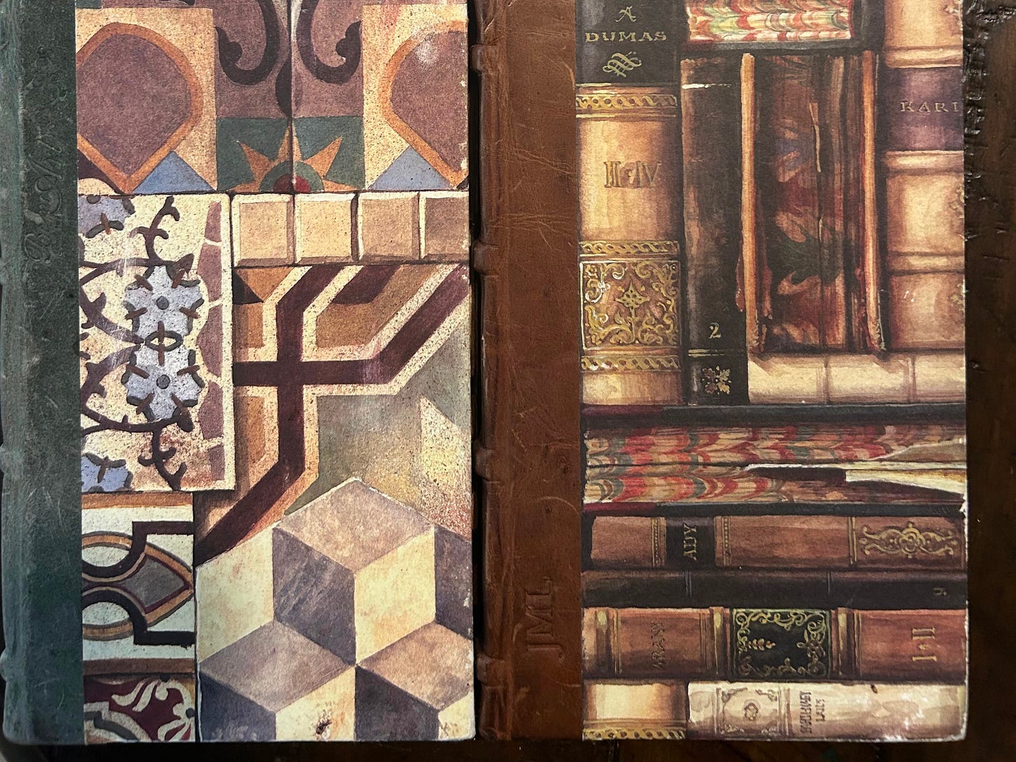 Two hardbound notebooks resting side by side. Each has leather binding on the left side; one of them has the initials JML inscribed in the leather. One has a series of geometric patterns on the cover; the other pattern is a stack of ancient books.