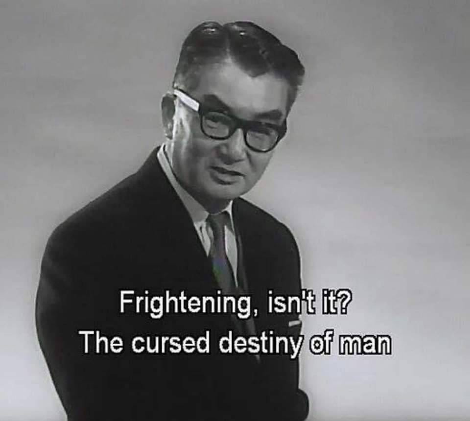 A man in a suit an glasses looking at the camera and saying "Frightening, isn't it? The cursed destiny of man."