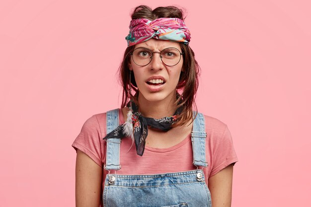 Young white woman wearing huge round glasses, a pink patterned scarf round her forehead that makes her brown hair stick up, denim overalls, a mismatching scarf round her neck, uneven short hair, and an expression that scrunches up her face and puts lines in her forehead. She is intended to look ugly.