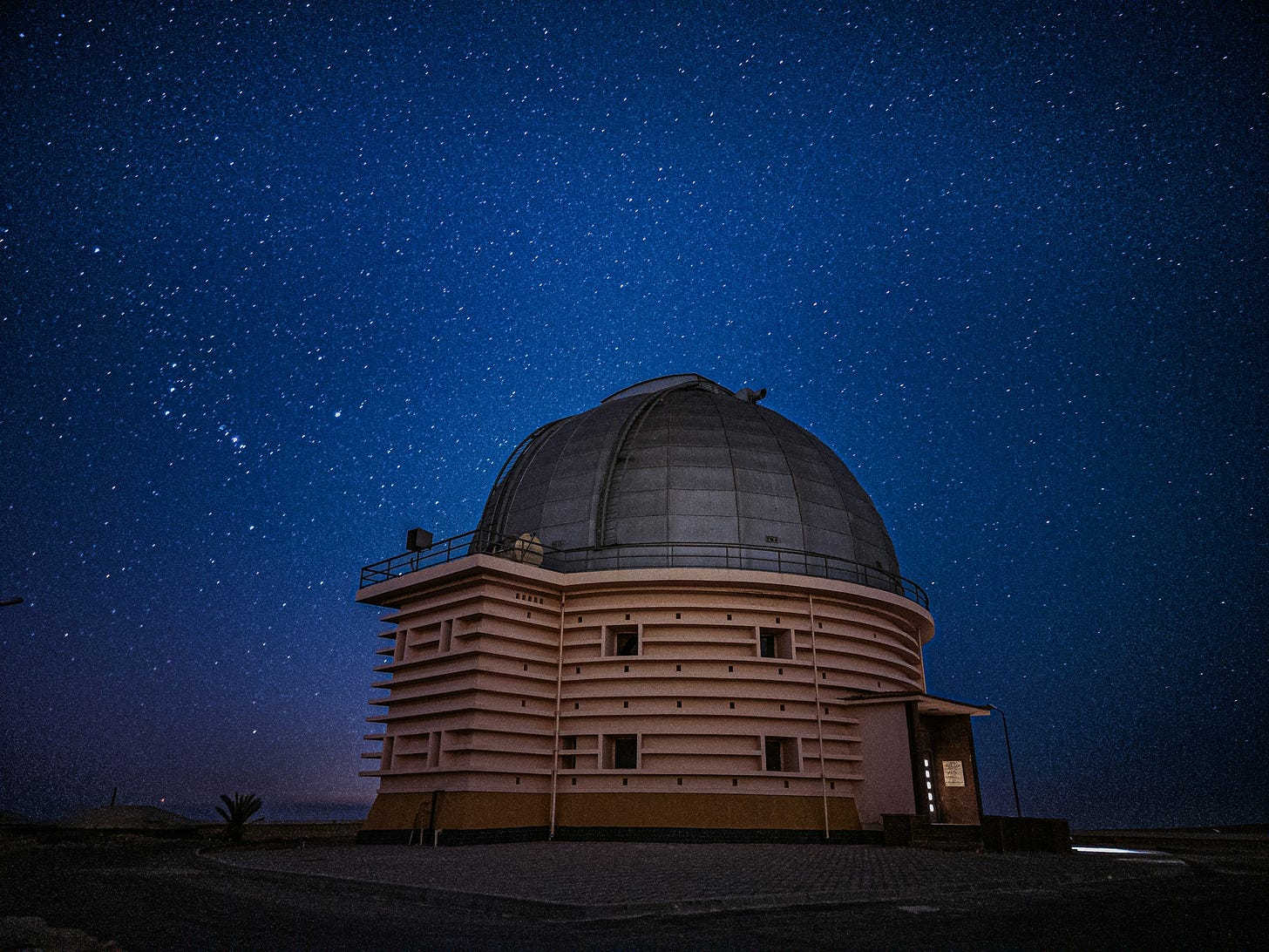 Cielo notturno pieno di stelle con l'edificio di un osservatorio astronomico al centro, sormontato da una cupola grigio scuro.