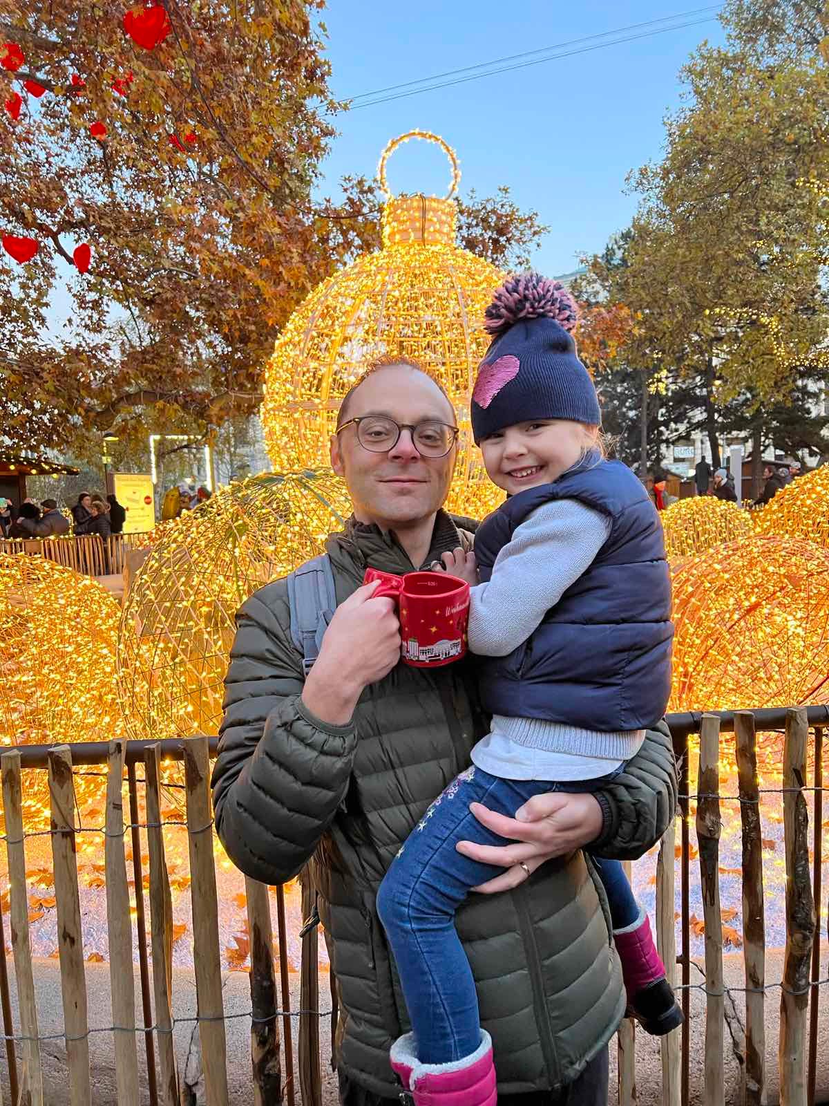 A man holds his young daughter and mug of mulled wine at Christmas market.