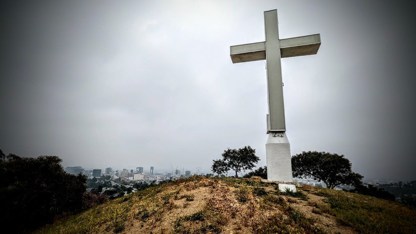 A daytime photo of the illuminated Hollywood Cross in Cahuenga Pass, with DTLA in the background