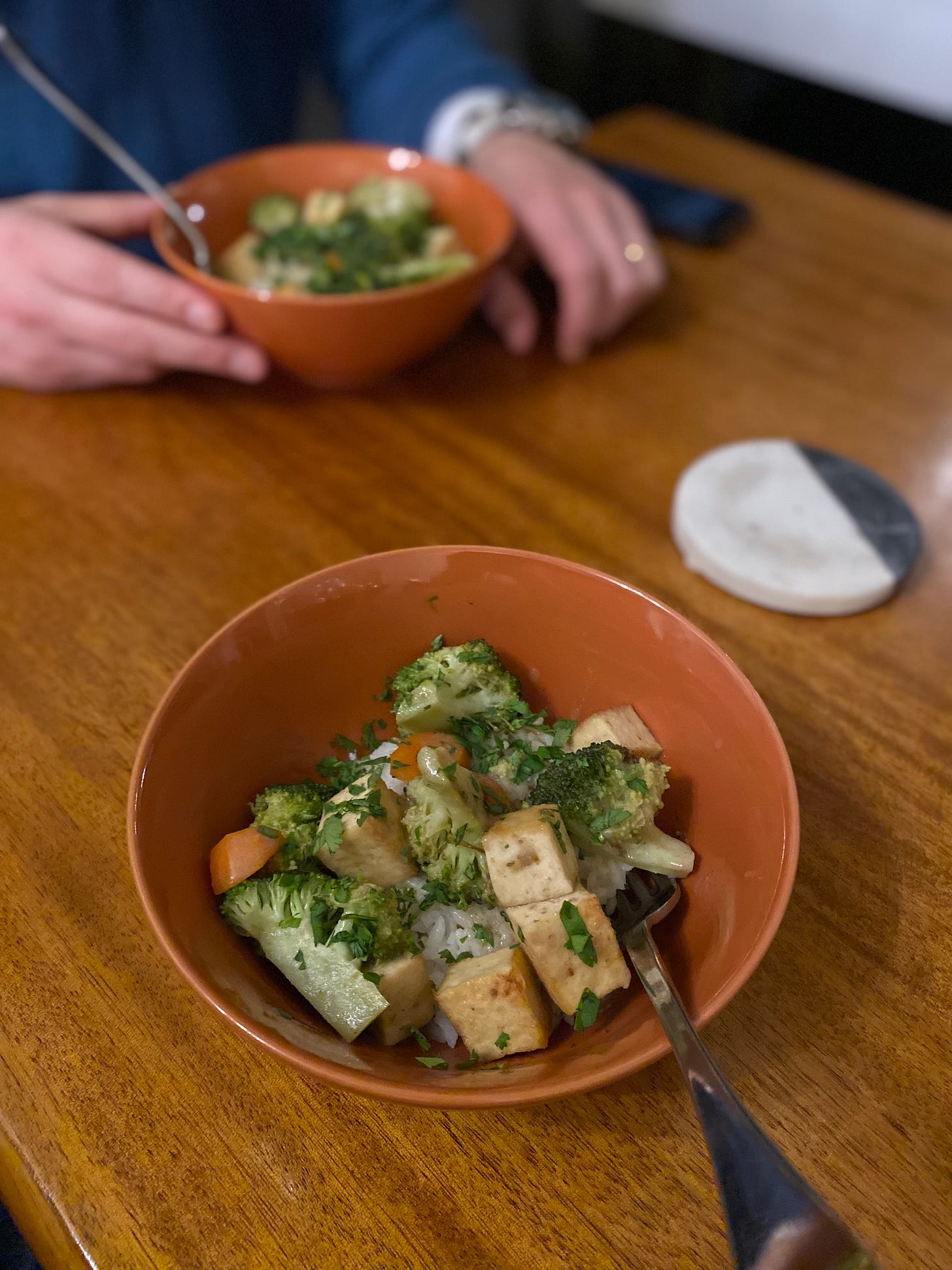 The curry described above, with cubes of tofu, broccoli florets, and carrot slices over rice. Everything is topped with chopped cilantro. Jeff's hand is touching his bowl of the same across the table.