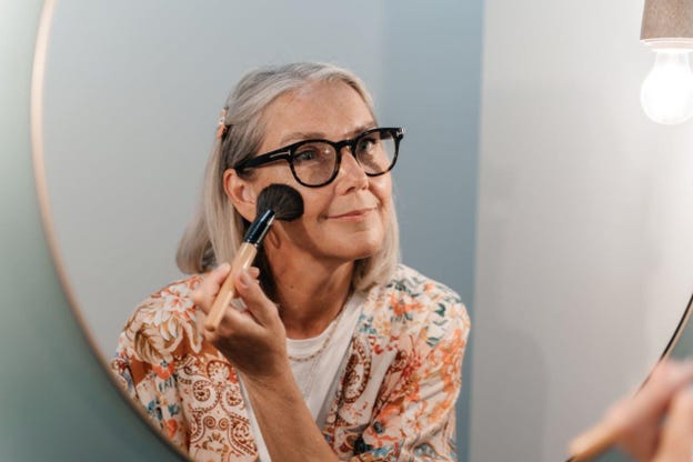 older woman with black-rimmed glasses and gray hair applying blush while looking into the mirror