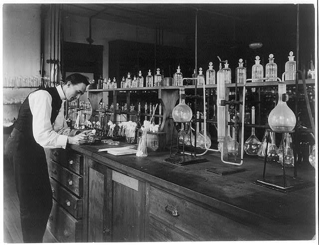 black and white photo of a man standing infront of a table filled with beakers and chemical bottles