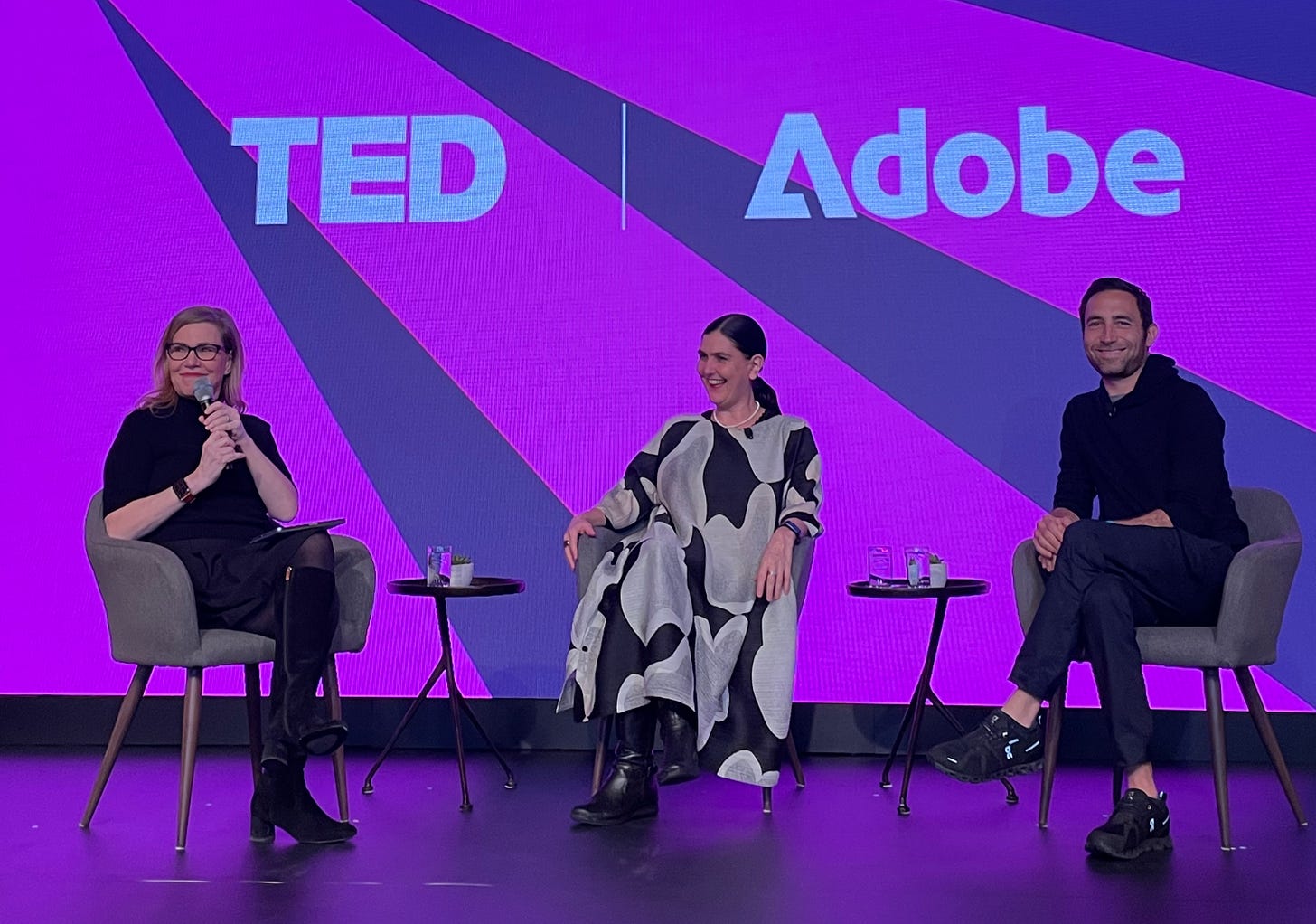 A photograph of three panelists seated on stage, from left to right: Debbie Millman, Honor Harger, and Scott Belsky, who smiles at the camera. The screen behind the speakers is a bold pattern of pink and navy with the text 'TED | Adobe'.