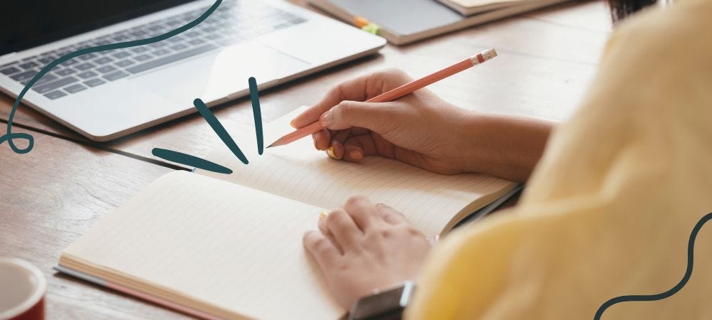 A person in a yellow jumper holds a pencil above a blank notebook with an open laptop in the background