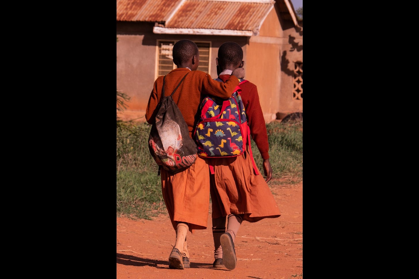 Two preteen African girls dressed identically in a red sweater and orange skirt walk home from school. One girl has her arm around the other's shoulders. The view is from the back.