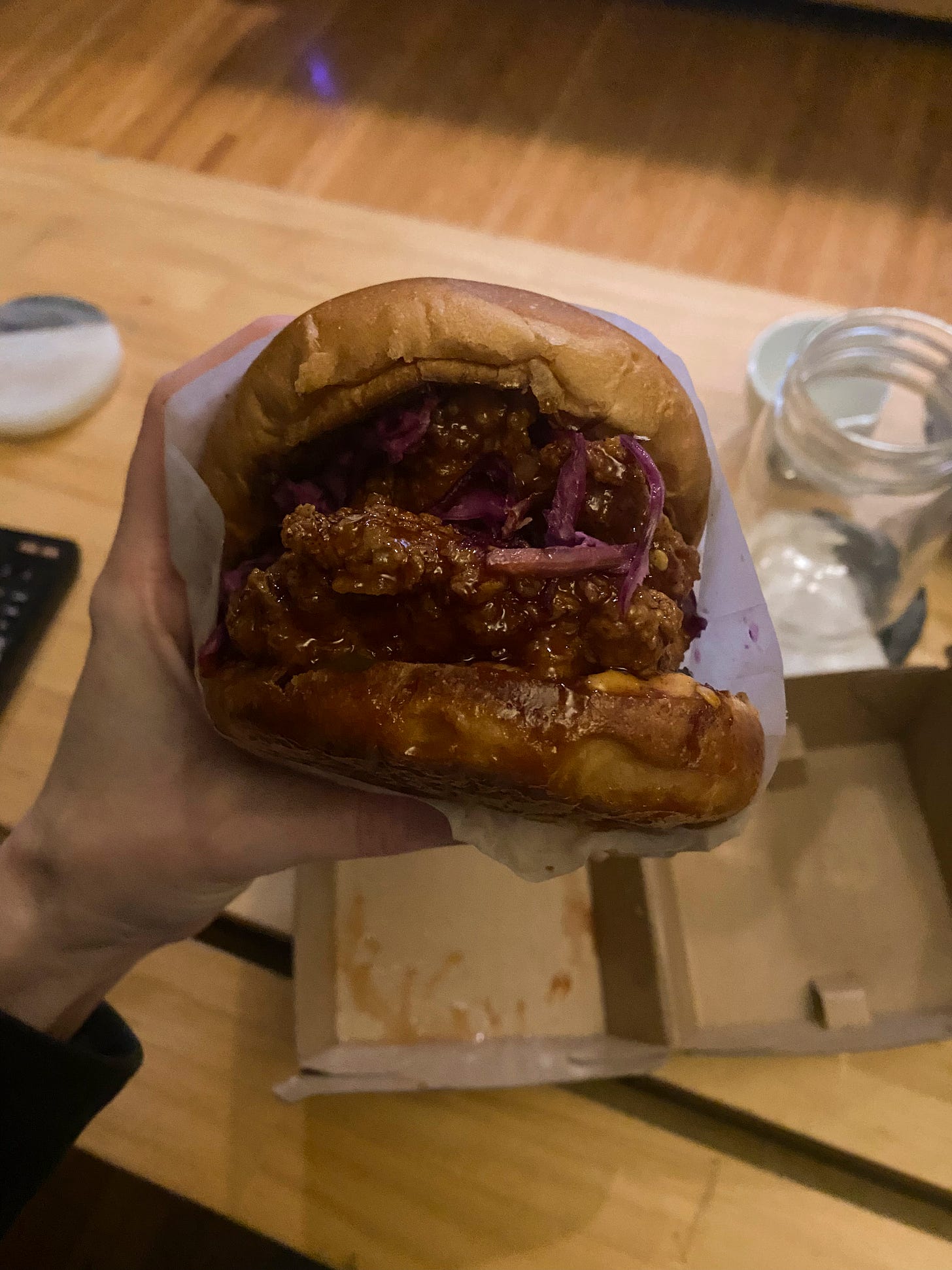 A messy-looking fried chicken sandwich in a reddish-brown sauce, with cheese and red cabbage visible at the edges. I'm holding it over top of the paper box it came on, which is sitting on the coffee table next to an empty glass on a coaster.