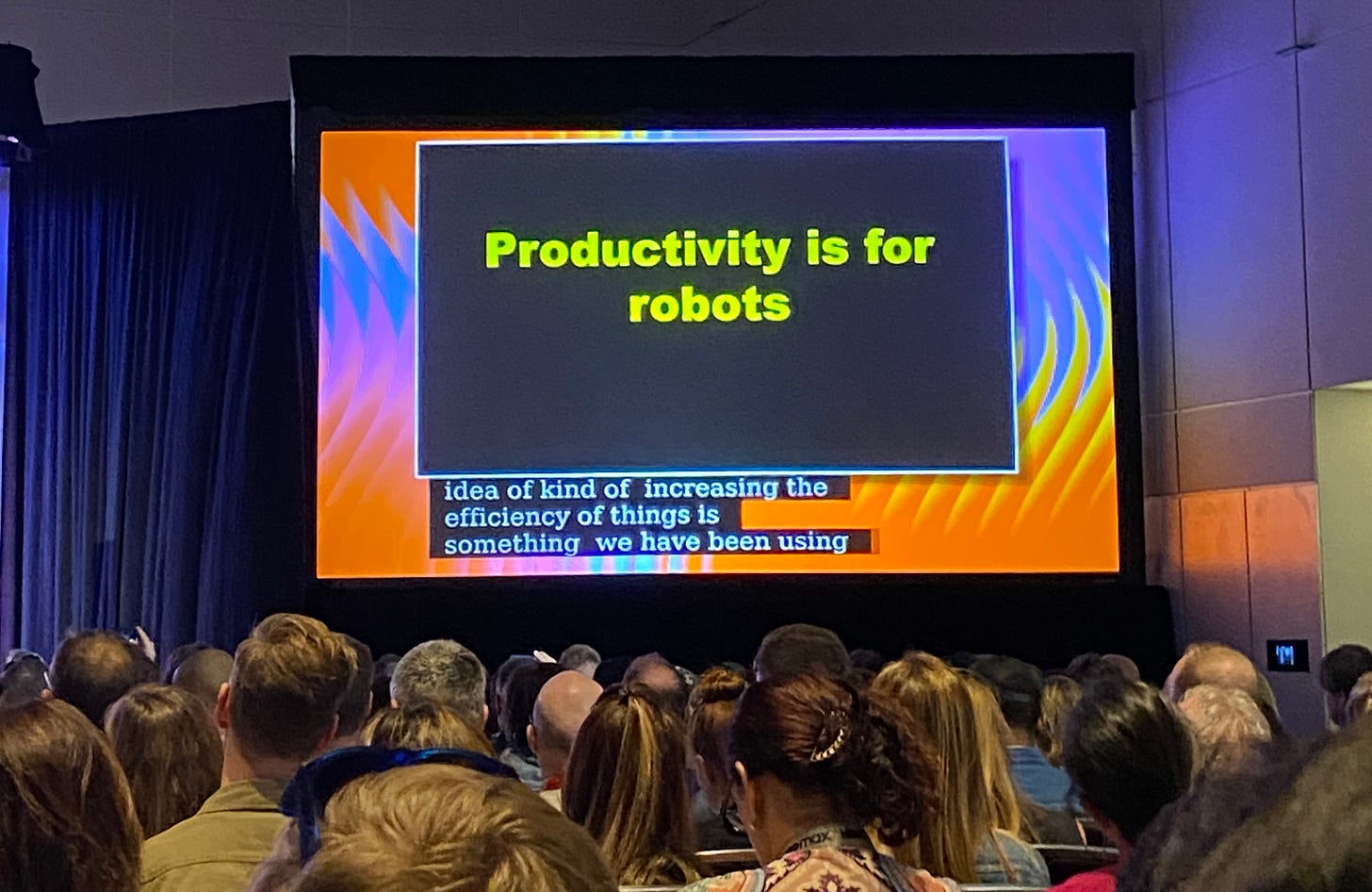 Imagem com uma plateia de costas e um slide sendo apresentado onde está escrito em inglês "Produtividade é para robôs"