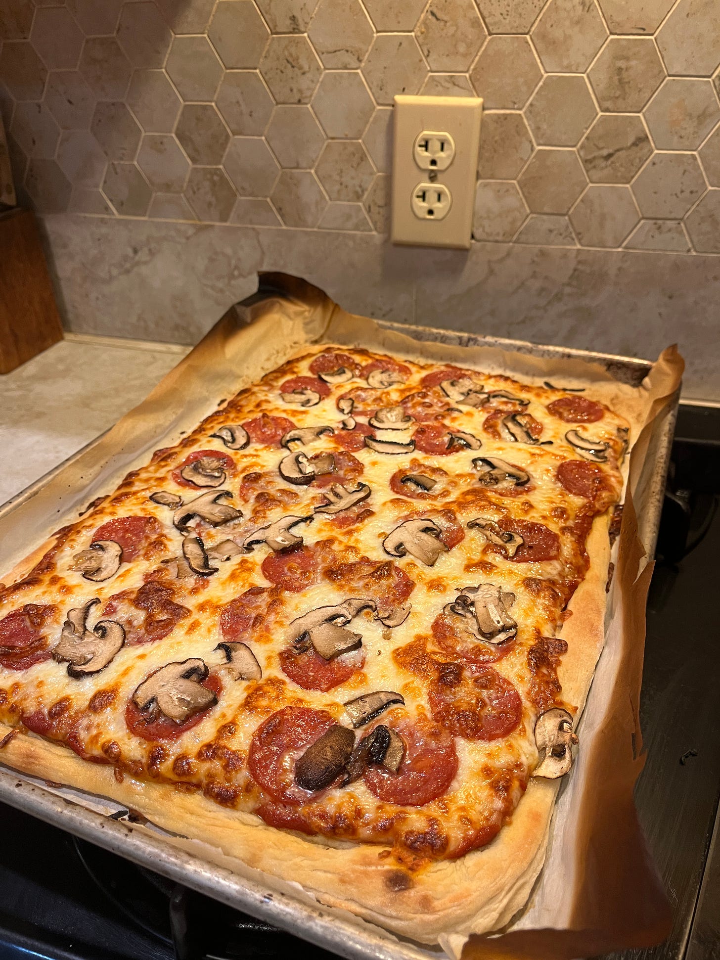 A rectangular pizza with salami and mushrooms sits on a slightly burnt parchment paper on a baking sheet