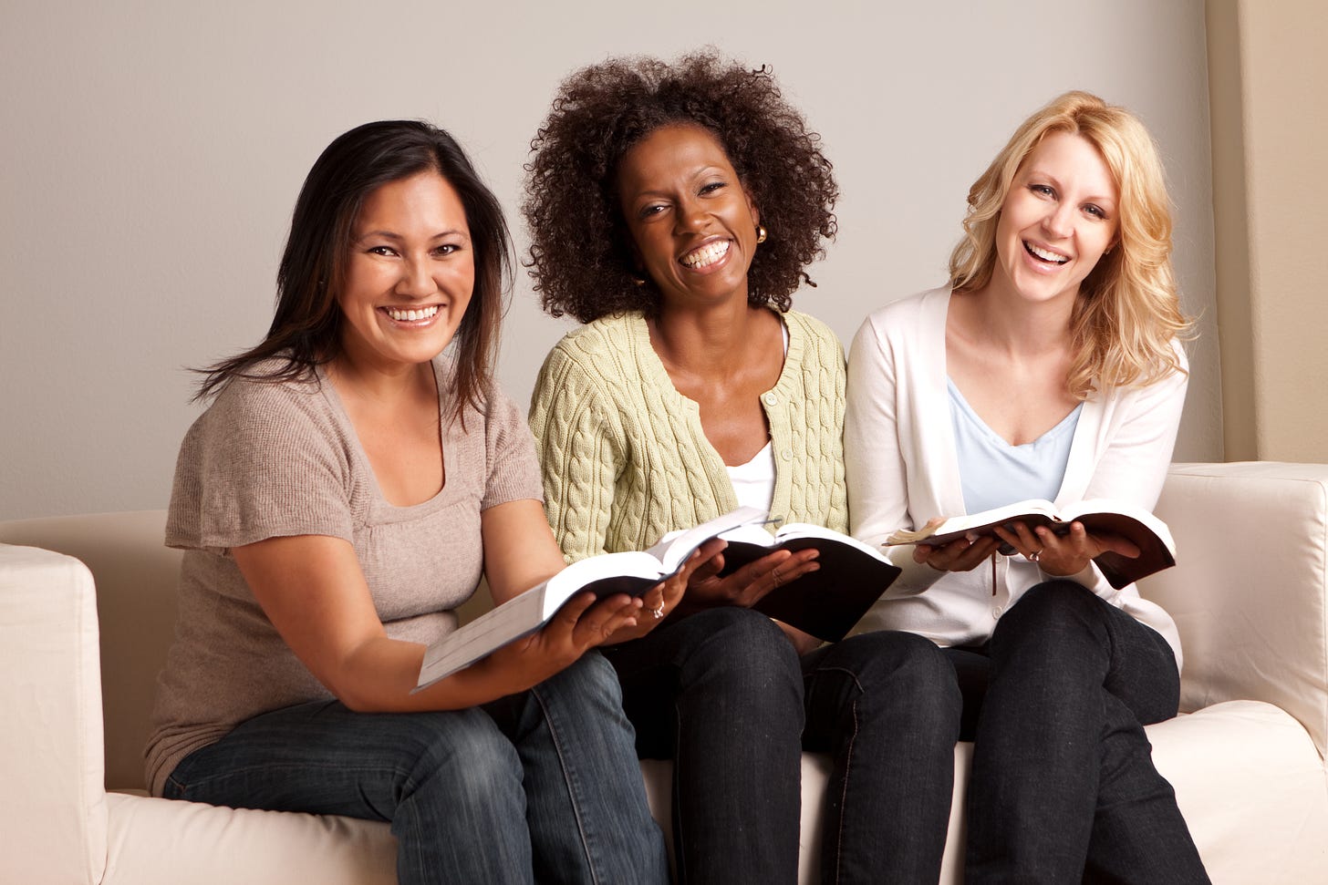 three smiling women sitting on a white coach all holding Bibles. They are from all different ethnicities, but they REALLY like the Bible