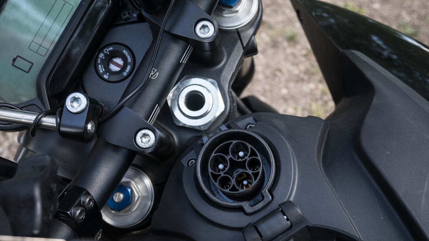 Close up photo looking down upon the front portion of the motorcycle, including the LCD dashboard, key ignition, handlebars and fork top, and the front section of the tank area, including an EV charging socket.