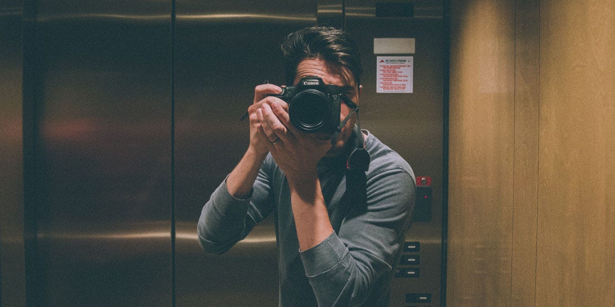 Foto colorida dentro de um elevador que mostra um homem caucasiano tirando uma foto no espelho.