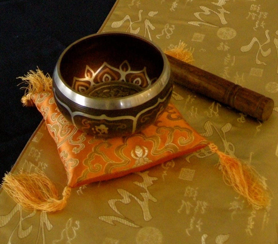 A small singing bowl resting on an orange cushion with the wooden mallet near it, all on a yellow silken runner