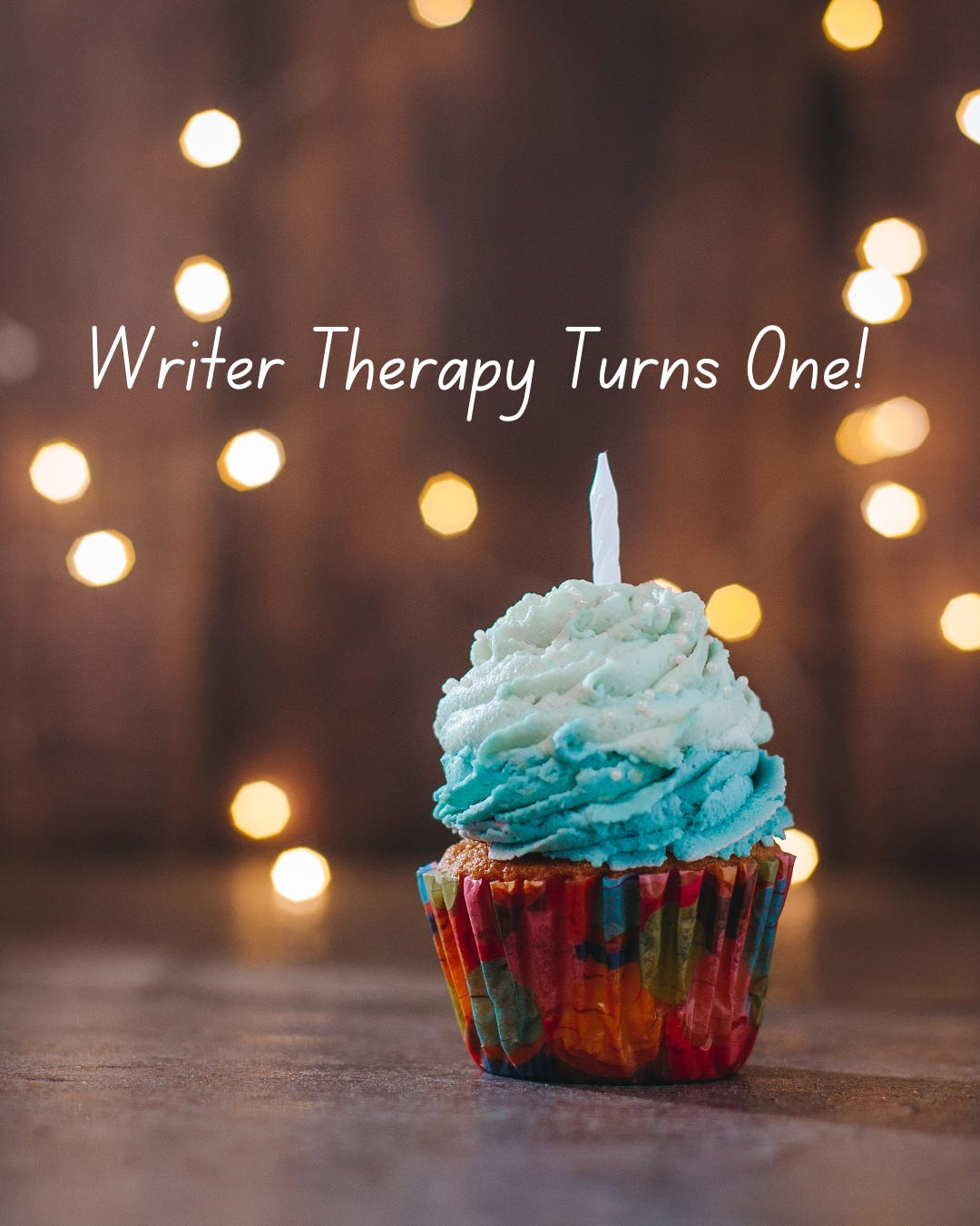 Stock image of a rainbow cupcake with a pile of icing in a gradient blue, and one candle. Twinkly lights in the background. Text reads, Writer Therapy Turns One!