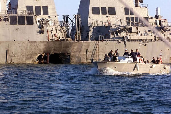 A large hole is seen in the side of a warship, as a smaller boat passes by with sailors aboard.