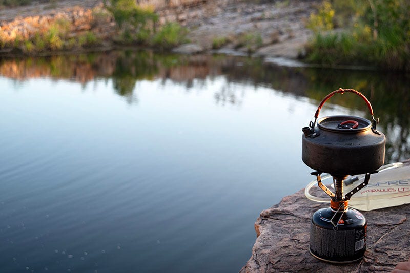 A teapot on a campstove overlooking a body of water.