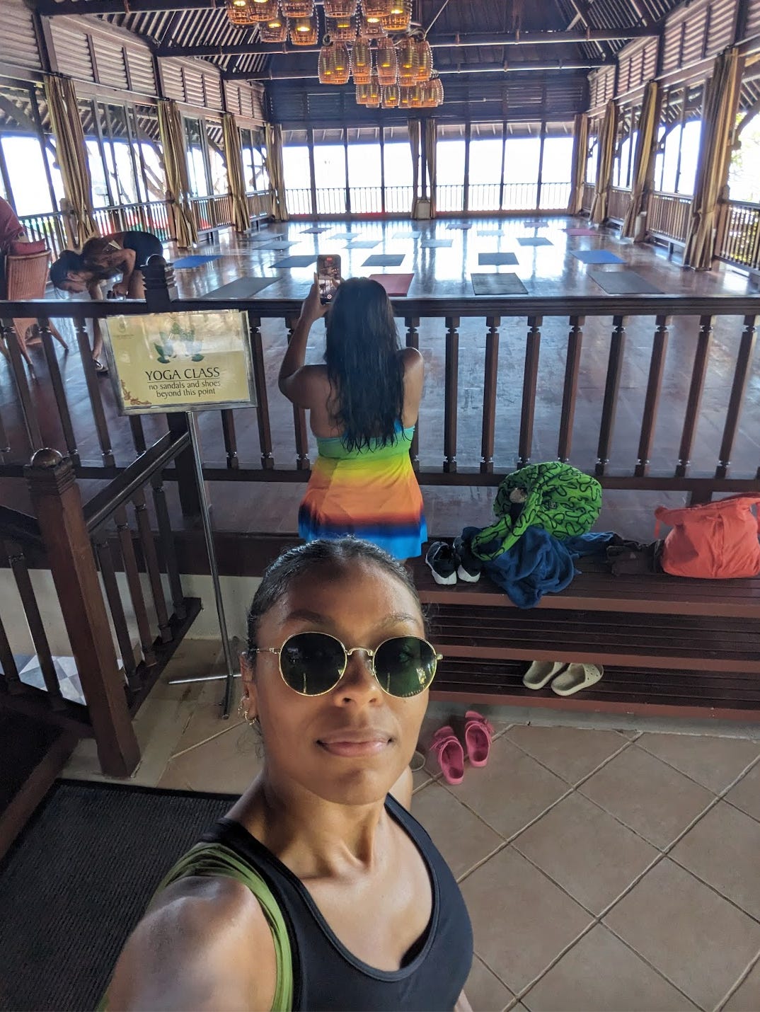 A selfie of Nathalie in front of a large yoga studio that has yoga mats lined up. There are large windows towards the front of the studio that overlooks the beach