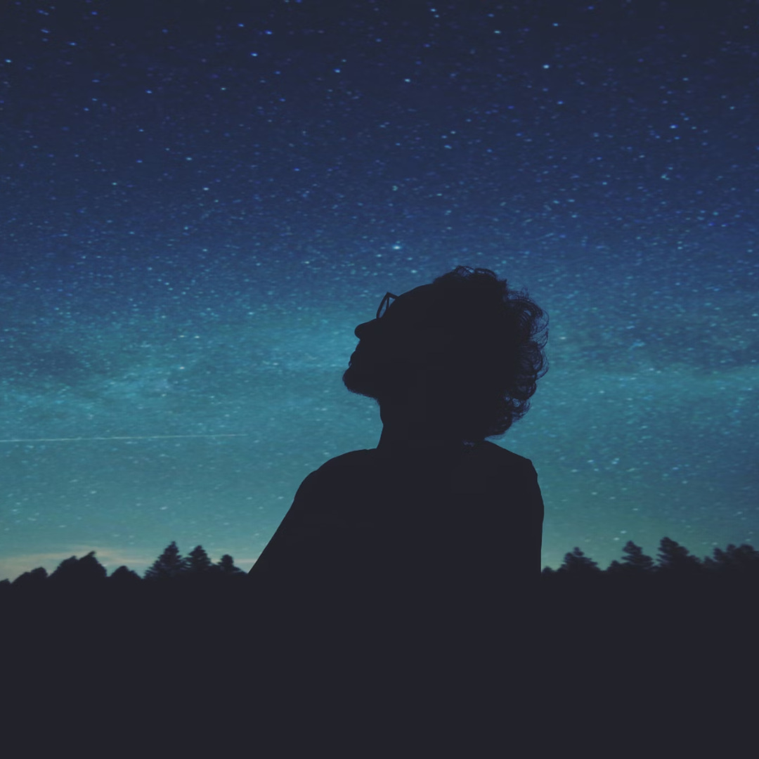 A man looks up at the milky way against a subtly glowing cool midnight blue sky.
