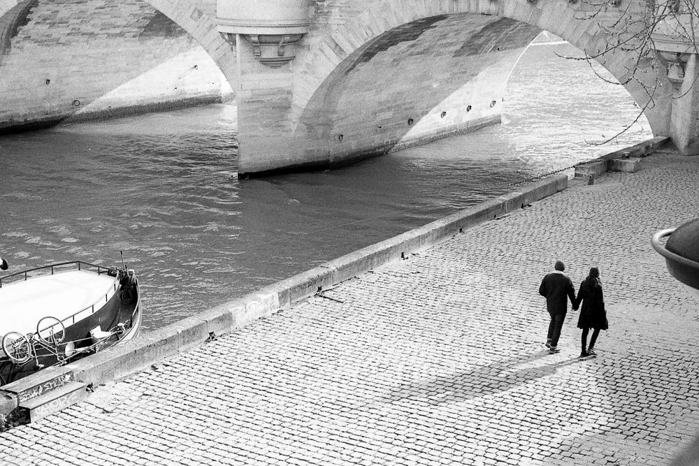 Black and white photograph of a couple walking hand-in-hand on a cobblestone promenade next to a river. They are walking towards the arches of a bridge and away from the prow of a boat