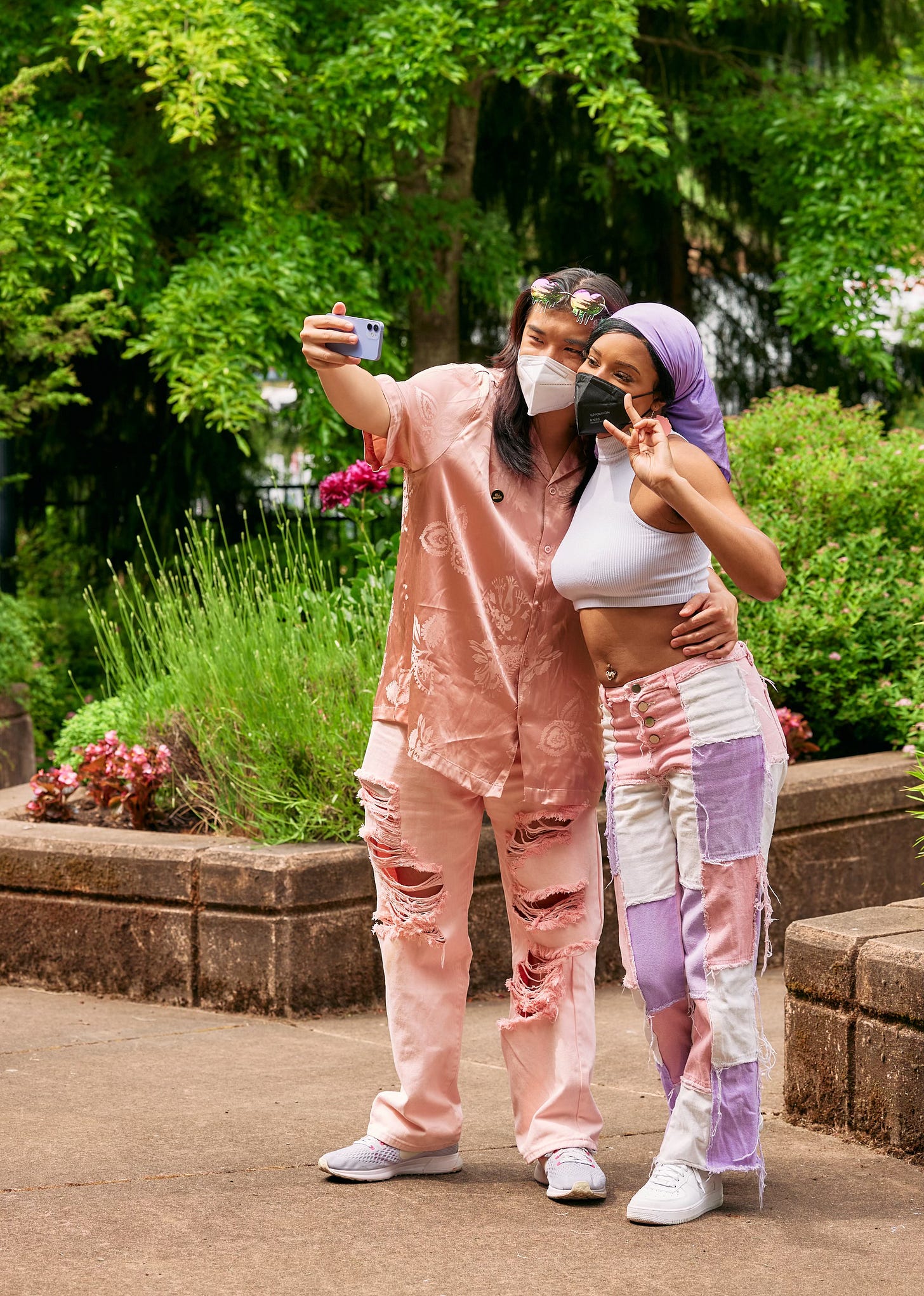 Portrait shot of a young masked autistic couple taking a selfie at a lush park with raised garden beds. On the left, the non-binary Asian person holds up a lavendar phone with one hand and wraps their free arm around their partner’s waist. On the right, the Black woman leans into her partner and poses with a peace sign. The couple has coordinating colors on, with the non-binary person in champagne pink, and the woman in lavender, white, and pastel pink.