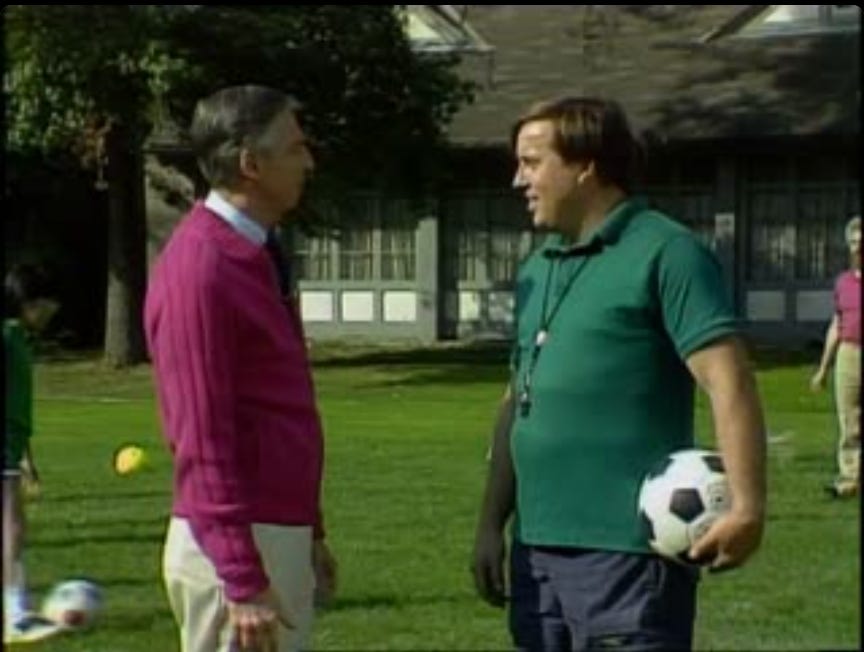 Mr Rogers and Dave Bartholomae, facing each other. They are standing on a soccer field and Dave is holding a soccer ball.