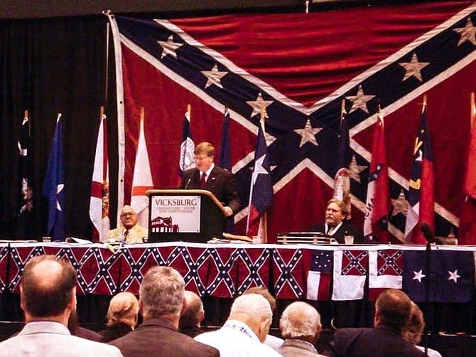 Mississippi governor Tate Reeves stands speaks at a podium in front of a massive Confederate flag a gathering 