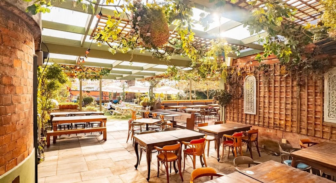 An outdoor dining location with wooden benches and seats, a green pergola, and brick walls, and plants around it An outdoor dining location with wooden benches and seats, a green pergola, and brick walls, and plants around it