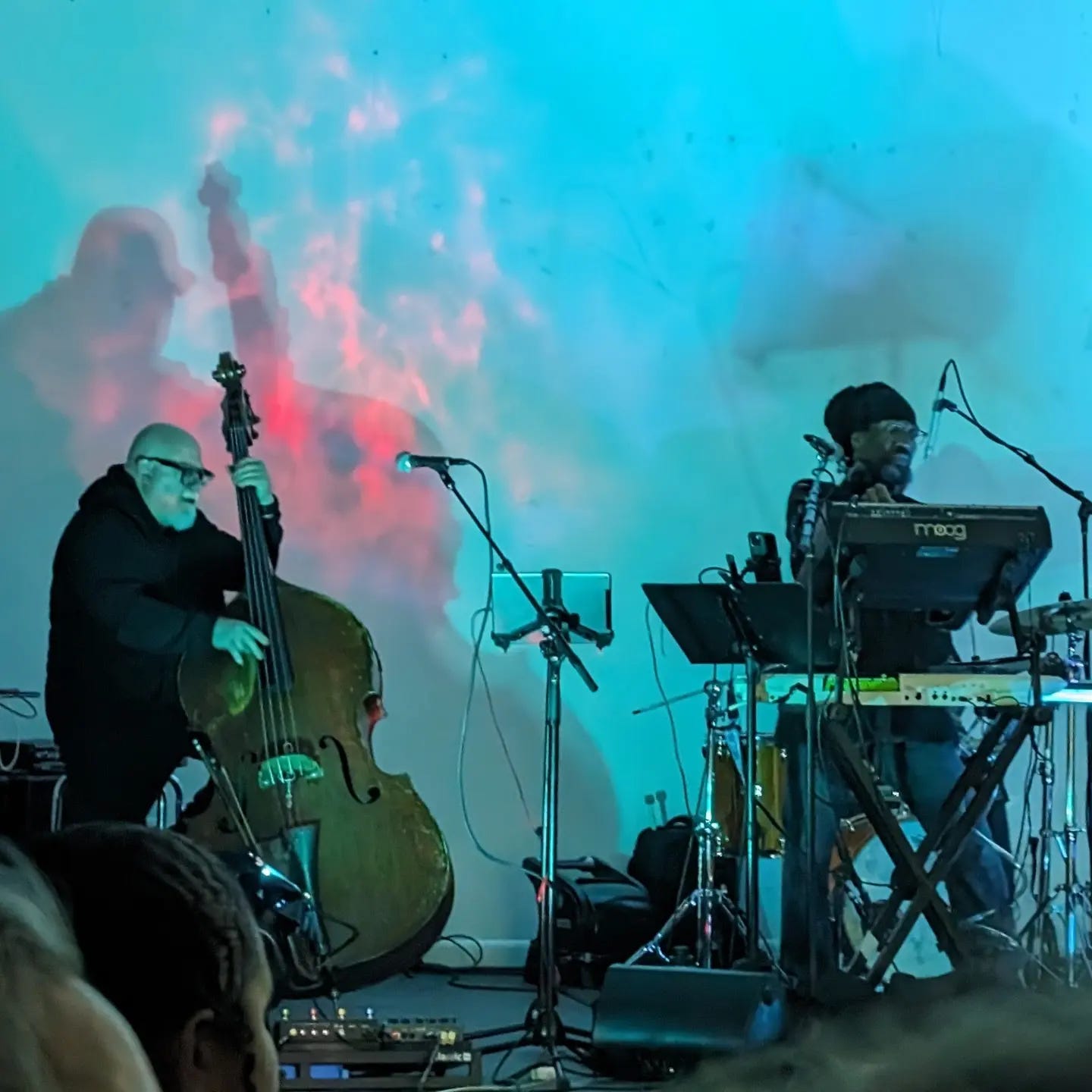 Phil Maneri playing upright bass, Jeffro Jam playing keys, drummer obscured, in front of blue and pink abstract projection