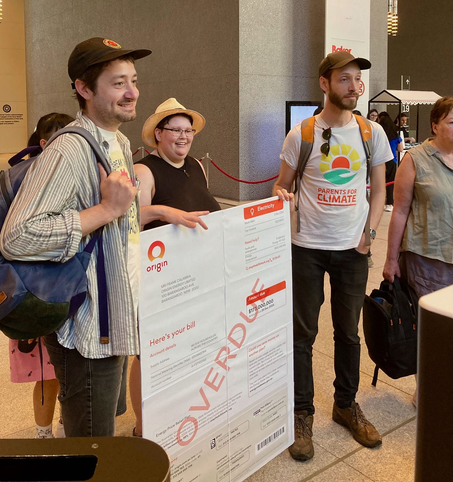 Three people standing in the foyer of an office building with a novelty sized bill. They are looking off camera towards Origin Energy representatives, who are not pictured. 